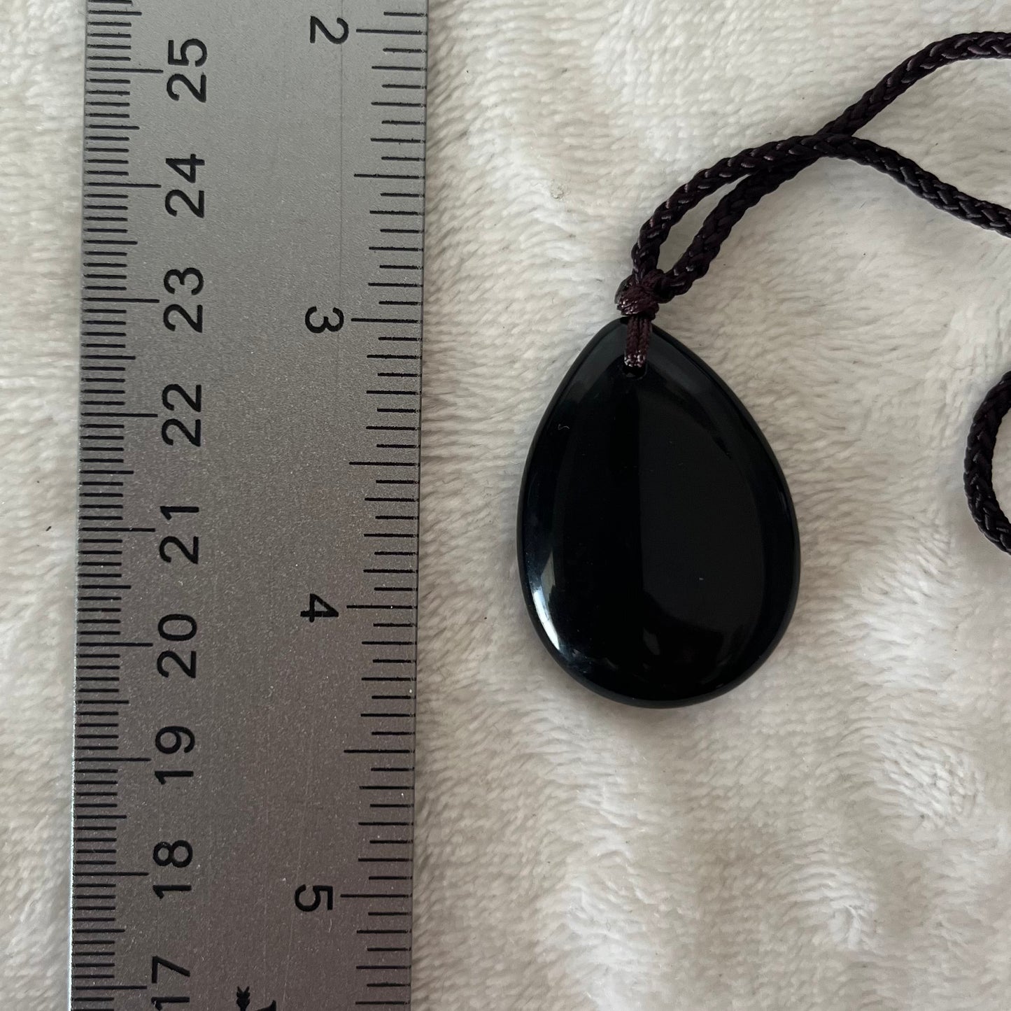 "Sleek obsidian teardrop pendant necklace, showcasing the beauty of black volcanic glass in a refined design, suspended from a delicated adjustable black cord displayed next to a ruler. Crystal pendant is approximately 1 1/4” long