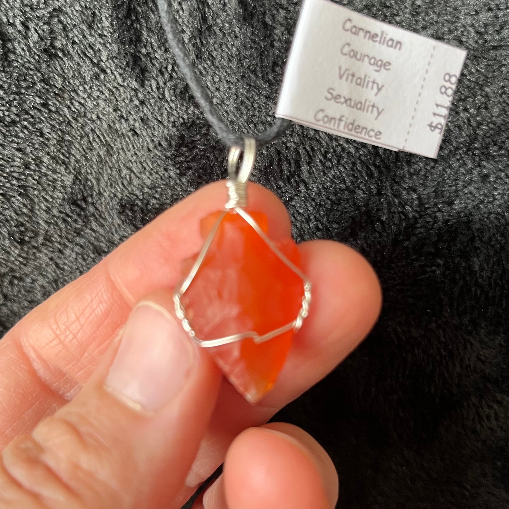 1 inch vibrant orange raw carnelian agate arrowhead, handmade, wire wrapped necklace, attatched to a 20 inch adjistable black cord, held delicately by three fingers.