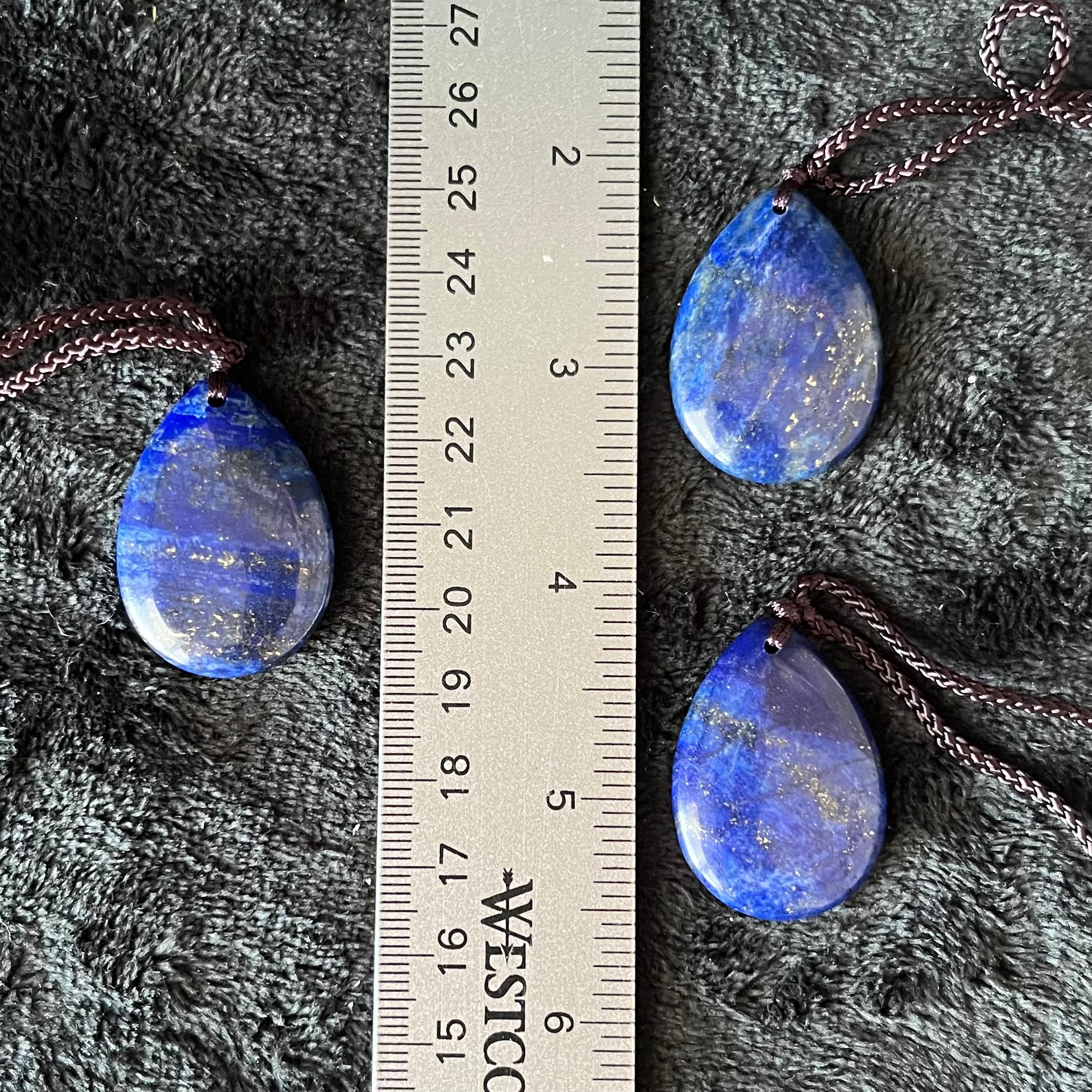 3 Exquisite lapis lazuli tear drop pendant necklaces, featuring deep blue gemstones suspended delicately from adjustable brown cords, displayed next to a ruler.  Pendants are approximately 1” long.