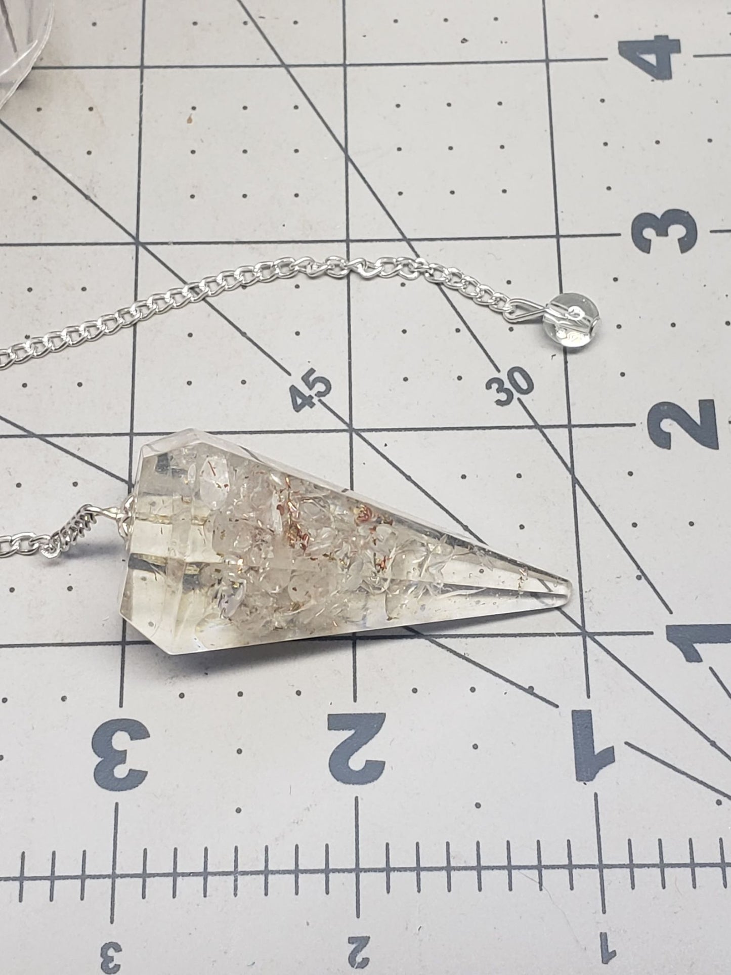 Clear Quartz 2 inch orgonite pendulum suspended from a silver 8 inch chain displayed next to a ruler
