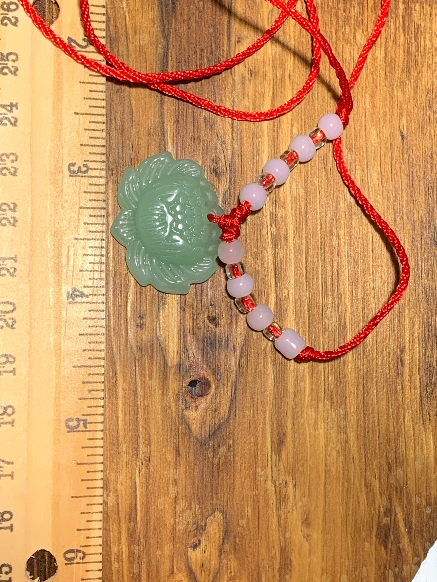 This green jade lotus pendant, carved with intricate detail, is attatched to a red cord with 4 white beads on either side, displayed next to a ruler to show size.