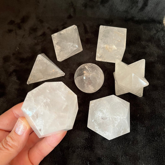 Seven-piece sacred geometry quartz set featuring intricate patterns and precision-cut designs for spiritual and aesthetic significance, Including a pyramid, cube, Merkaba, sphere, dodecahedron, decahedron, and octahedron.