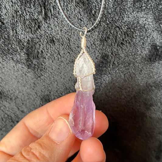 purple and white dragon tooth amethyst silver wire wrapped pendant attatched to a black cord, approximately 2 inches long, held delicately by 3 fingers against a black background