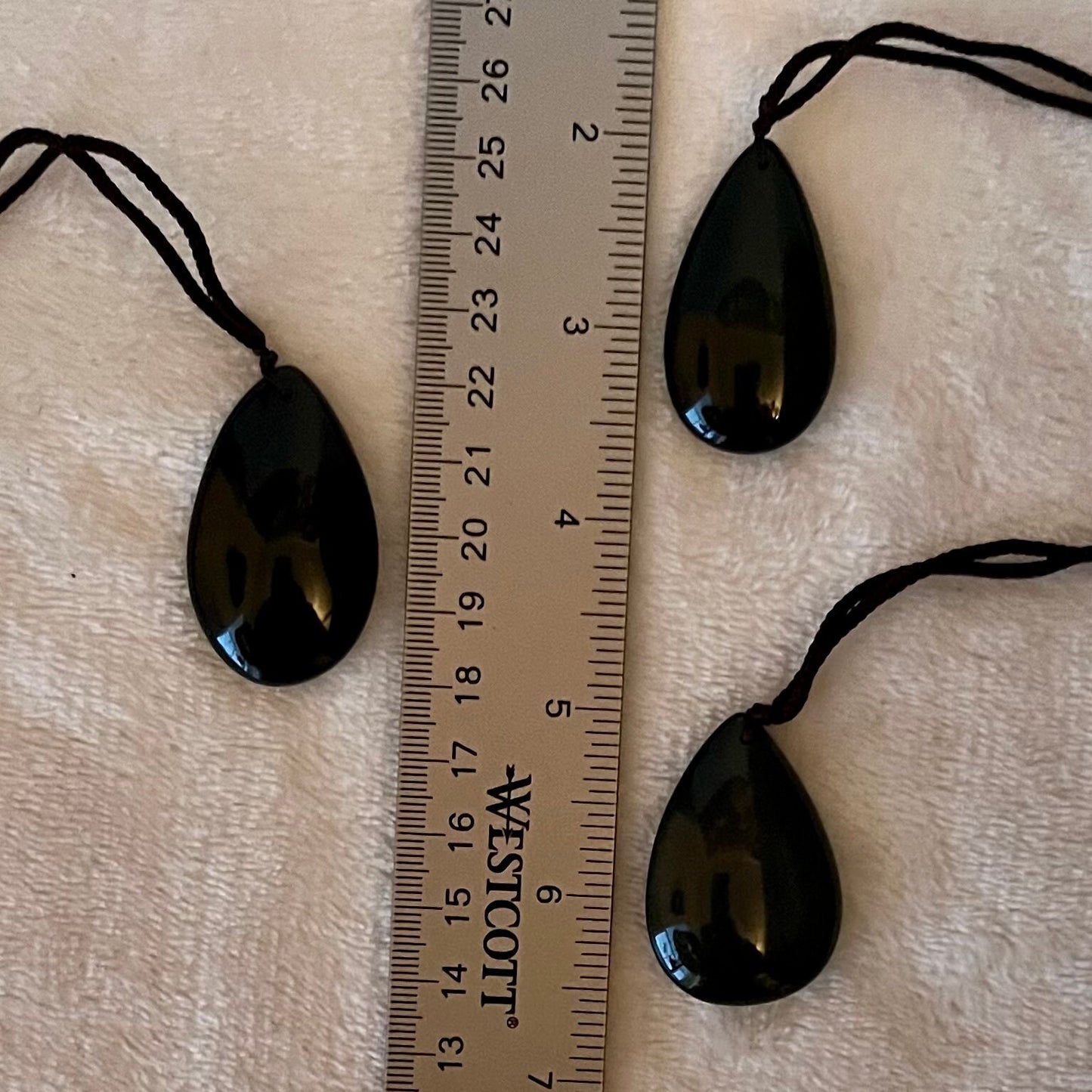 3 Iridescent rainbow obsidian tear drop pendant necklaces, showcasing a spectrum of captivating hues, gracefully hung from asjustable black cords and displayed next to a ruler to show size. Pendants are approximately 1 1/4” long