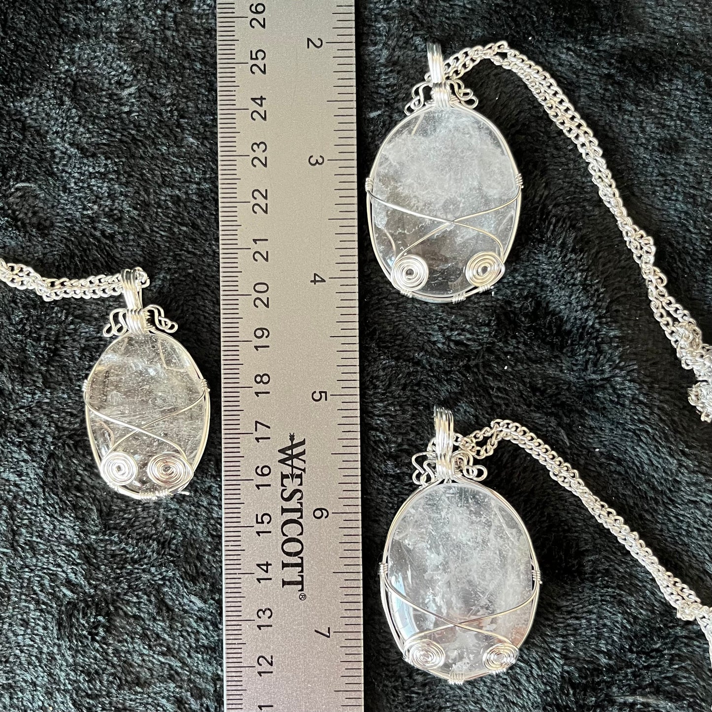 Three silver fancy wire wrapped clear quartz oval stone pendants attatched to a silver chains, displayed against a black background next to a ruler. eaxh oendant is 1 3/4" - 2 1/4" long