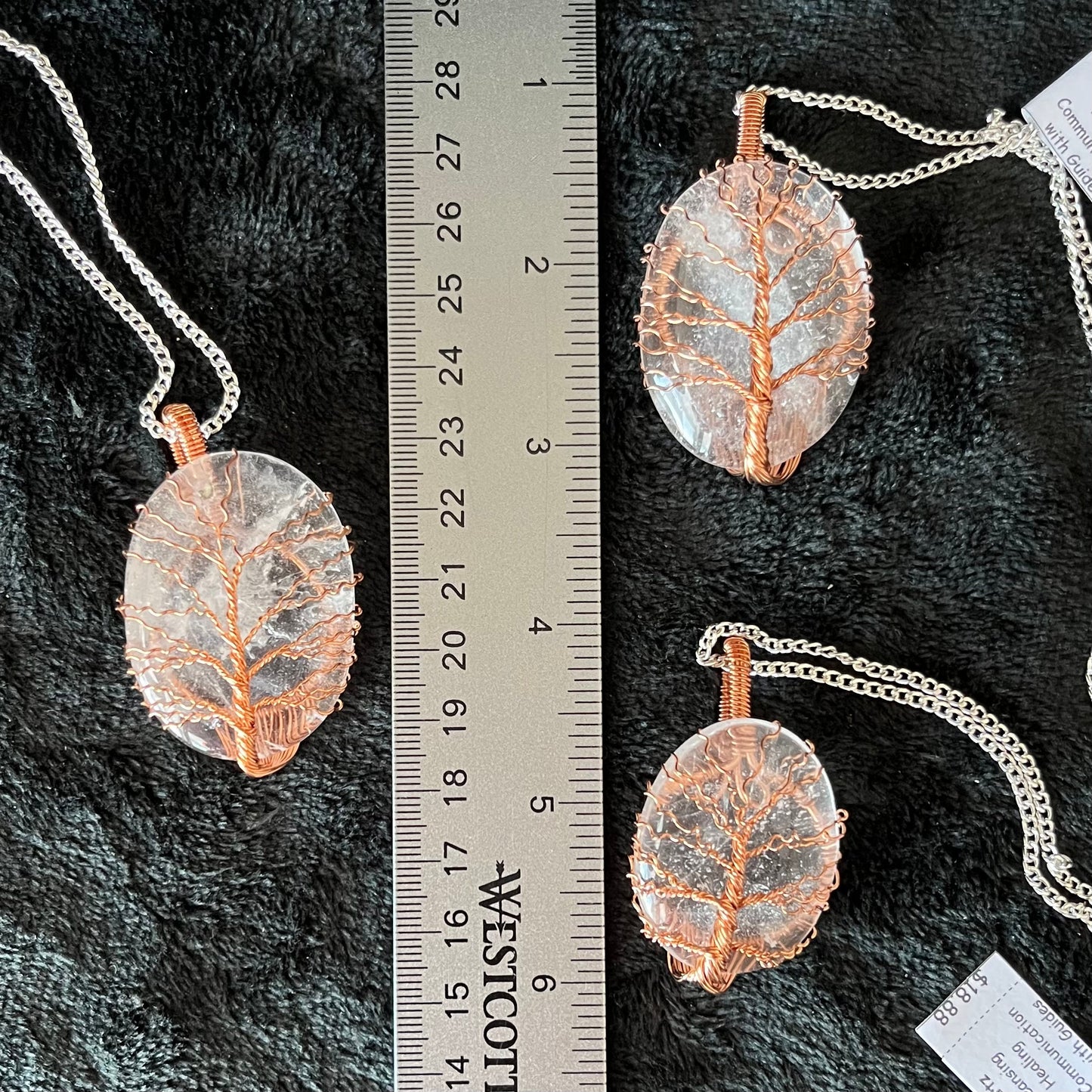 Three tree of life copper wire wrapped clear quartz oval stone pendants attatched to a silver chains, displayed against a black background next to a ruler. eaxh oendant is 1 3/4" - 2 " long