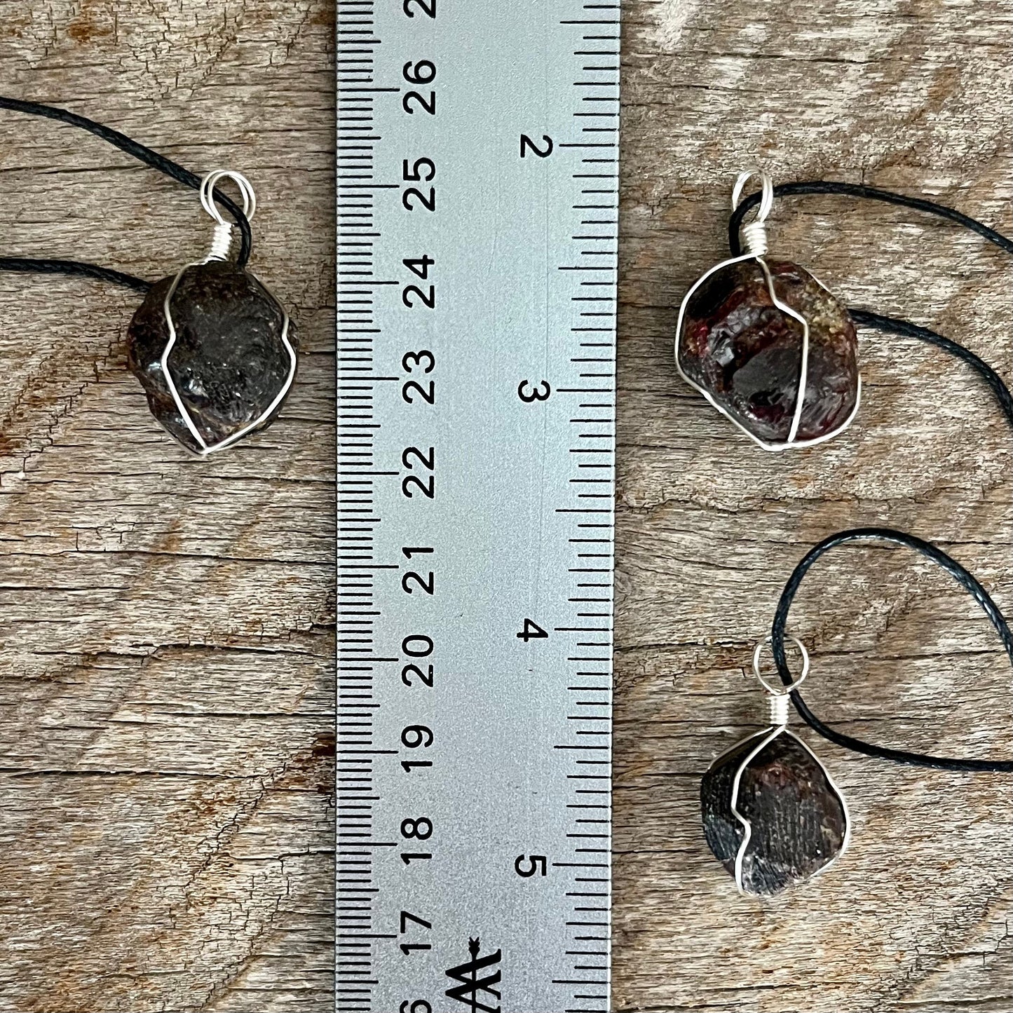 3 silver wire wrapped garnet crystal pendants attatched to adjistable black cords, next to a ruler. pendant vary in diameter from 1/2" to 3/4"