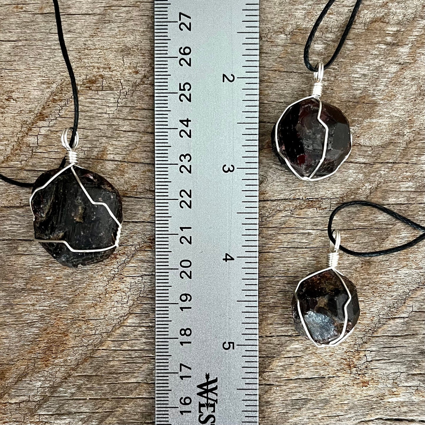 3 silver wire wrapped garnet crystal pendants attatched to adjistable black cords, displayed next to a ruler. pendant vary in diameter from 1/2" to 1"
