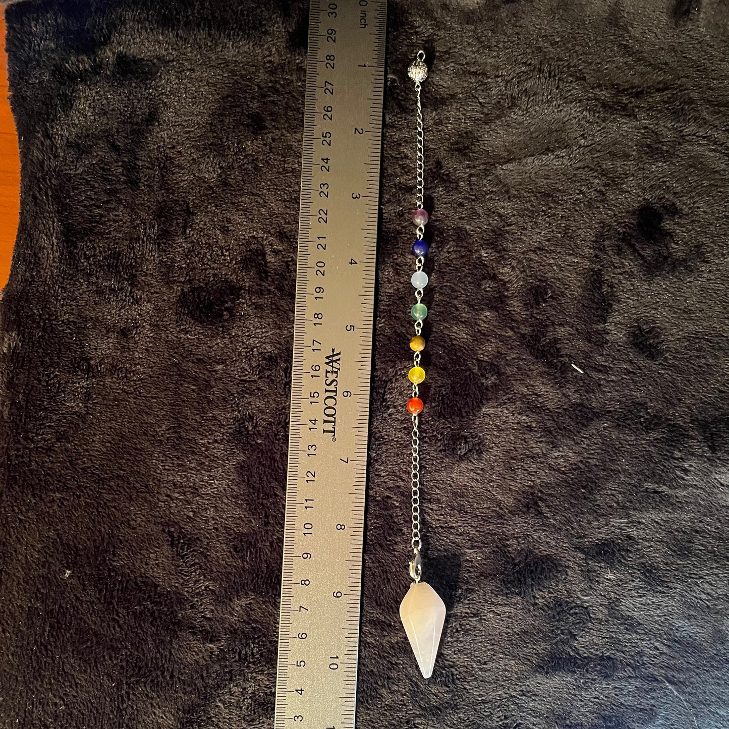 close up of rose quartz pendulum attatched to a silver chain containing chakra beads, next to a ruler to show 8 inch chain and 1 1/2 inch pendulum