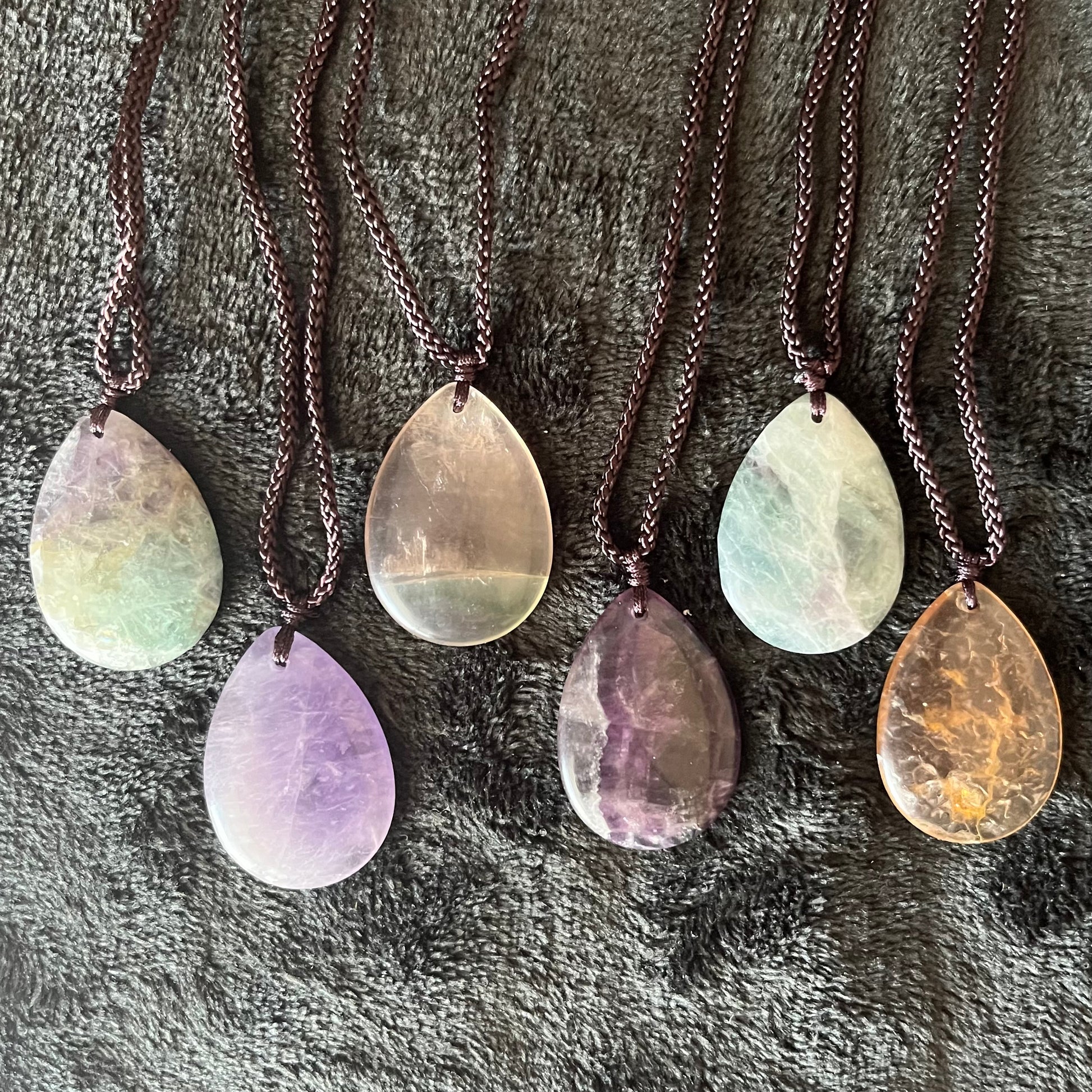 6 Captivating fluorite teardrop pendant necklaces, displaying a spectrum of vibrant colors, elegantly suspended from delicate asjustable black cords , adding a touch of enchantment to your jewelry collection."