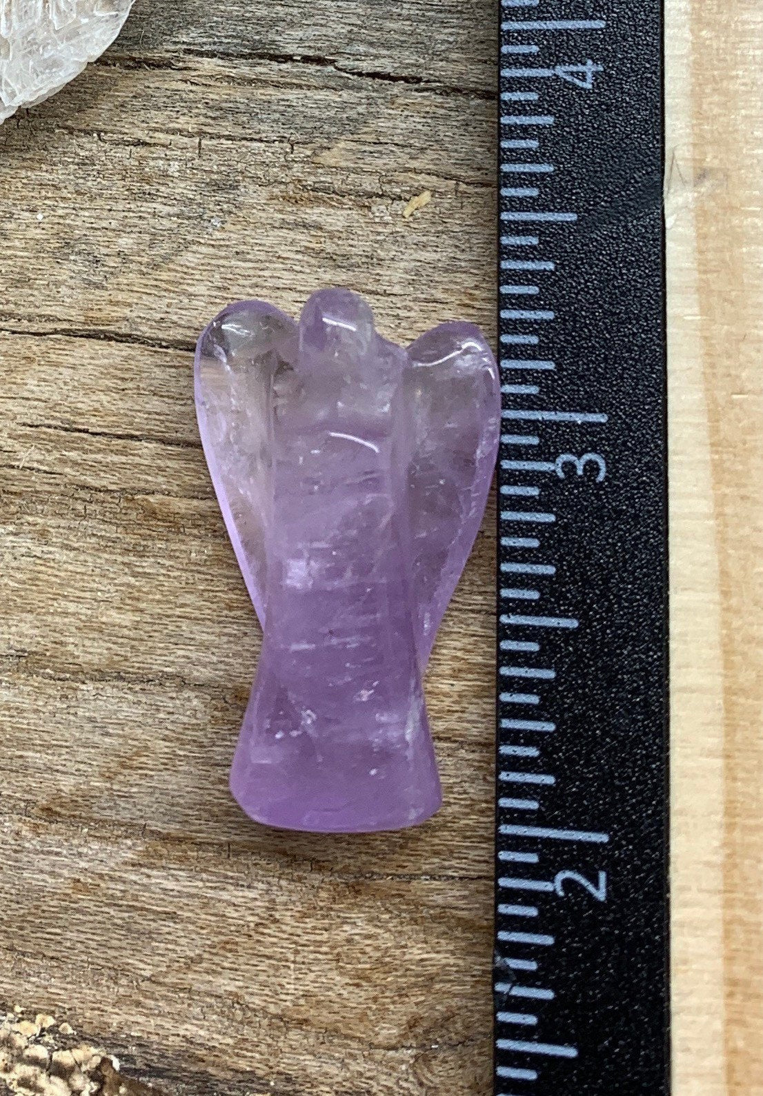 Small amethyst angel carving, featuring a serene and detailed design in translucent purple hues, displayed next to a ruler.  Amethyst Angel is approximately 1 1/4” tall.