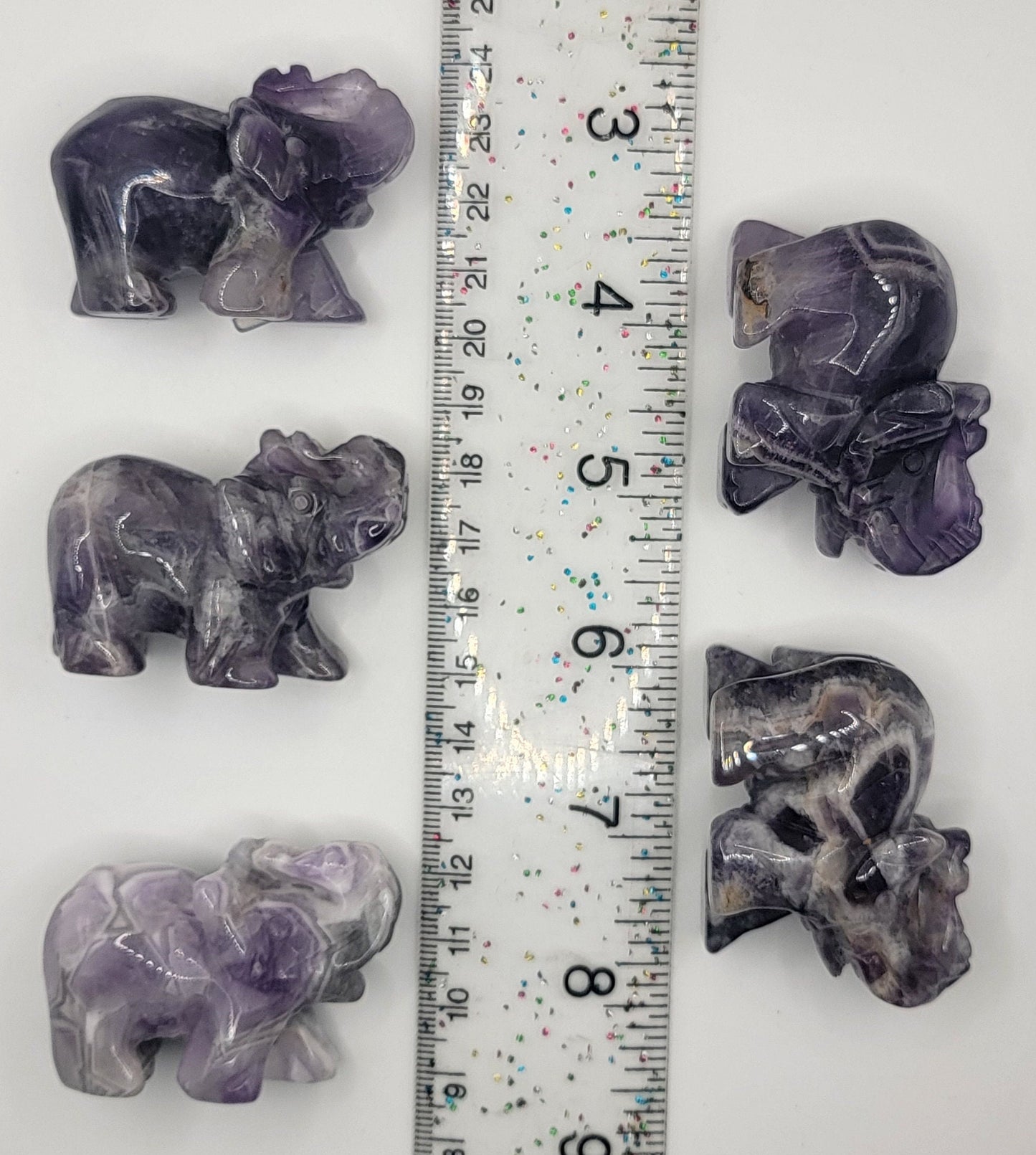 amethyst elephant carved figurines, showcasing intricately detailed craftsmanship and rich purple tones, displayed next to a ruler to show size. Amethyst elephants are approximately 2” x 1 1/2”.