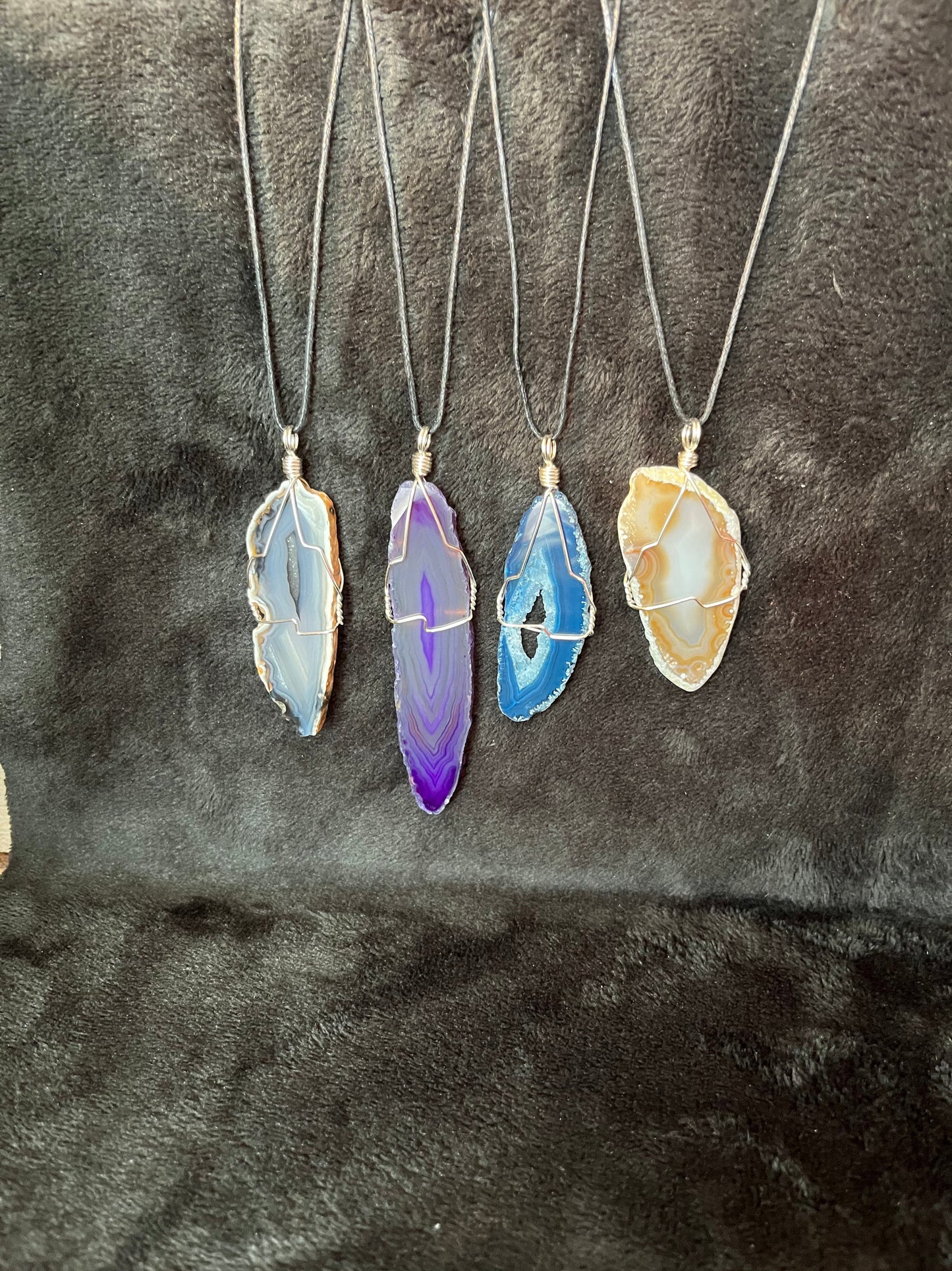 4 silver wire wrapped agate slices in purple, yellow, blue, and grey attached to adjistable black cords displayed against a black background to enhance their vibrant colors.  each necklace contains a tag with the crystal heaing properties listed including issom, growth, and psychic ability.