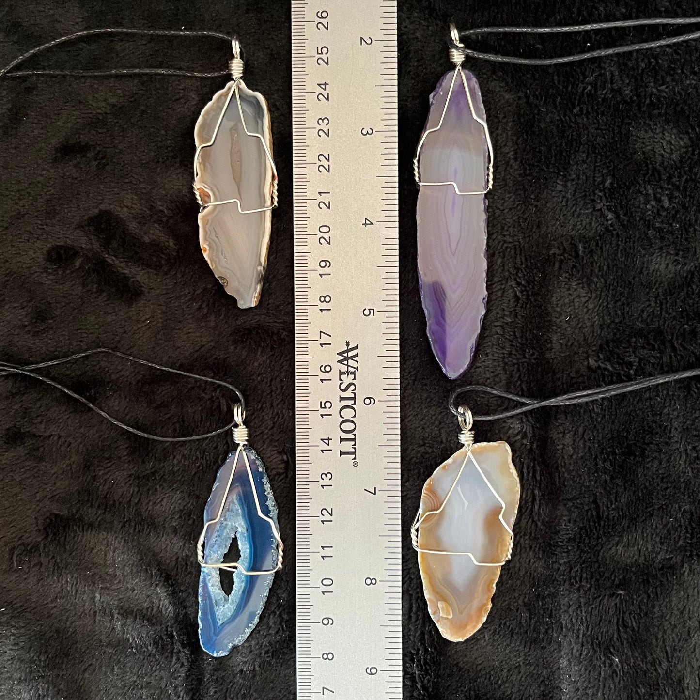 4 silver wire wrapped agate slices in purple, yellow, blue, and grey attached to adjistable black cords displayed against a black background to enhance their vibrant colors.  the Agate necklaces are displayed next to a ruler to show the size of each pendant.