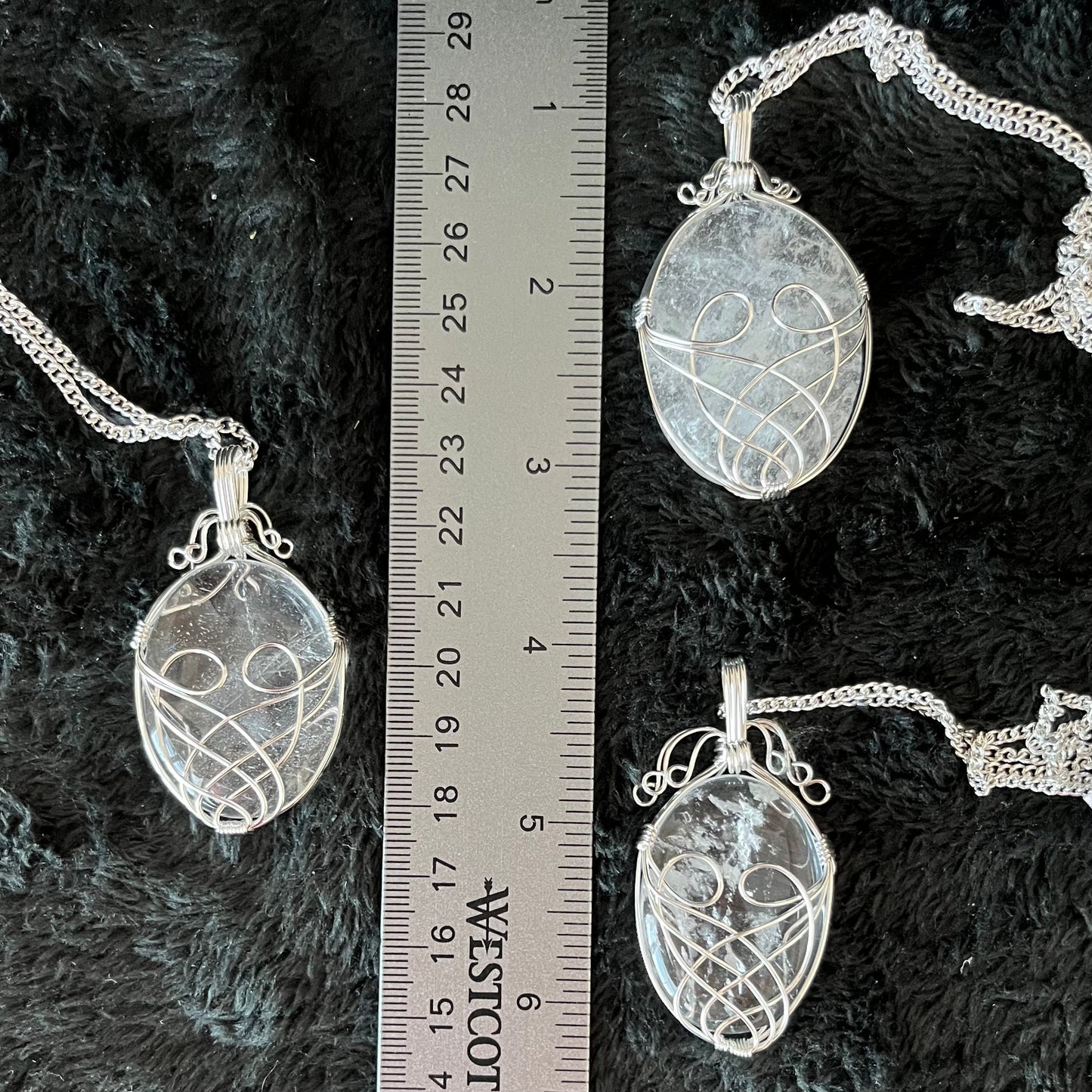 Three silver fancy wire wrapped clear quartz oval stone pendants attatched to silver chains, displayed next to a ruler.  quartz ovals are 1 3/4" - 2 1/4" long
