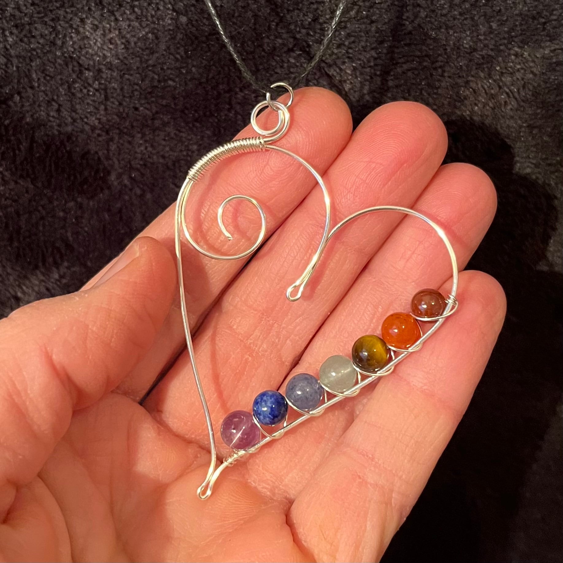 7 chakra beads spiral wire wrapped with silver wire in a line along the inside edge of a silver heart. attatched to a an adjustable black cord