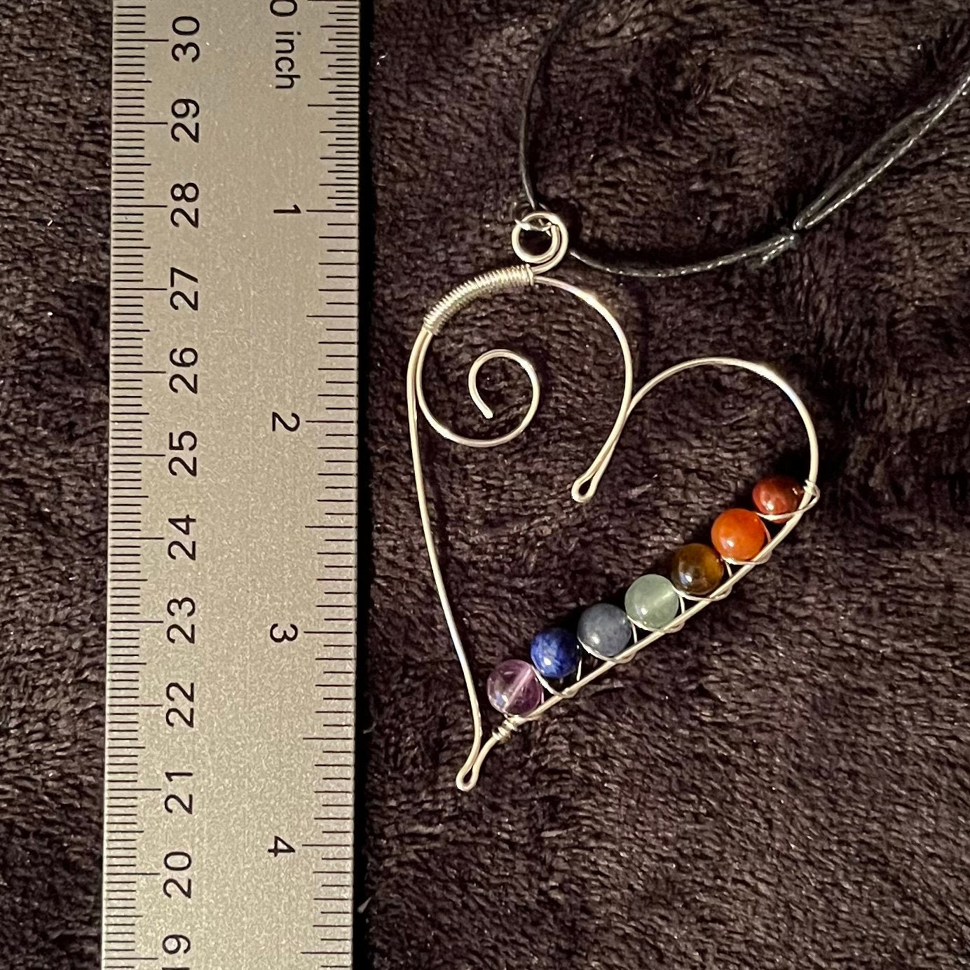 7 chakra beads spiral wire wrapped with silver wire in a line along the inside edge of a silver heart. attatched to a an adjustable black cord, displayed next to a ruler.  pendant is approximately 2 1/2 inches long.