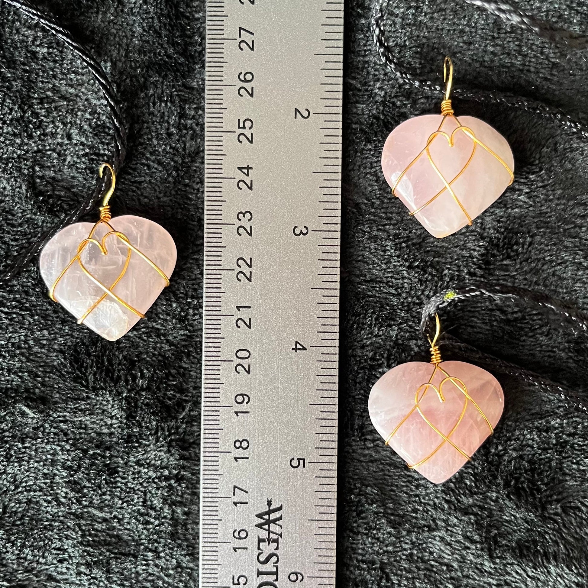 Three Wire-wrapped rose quartz heart necklaces: a gold wire intricately wraps around polished rose quartz heart pendants, hanging on a delicate chain, displayed next to a ruler.  hearts arw approximately 1" long