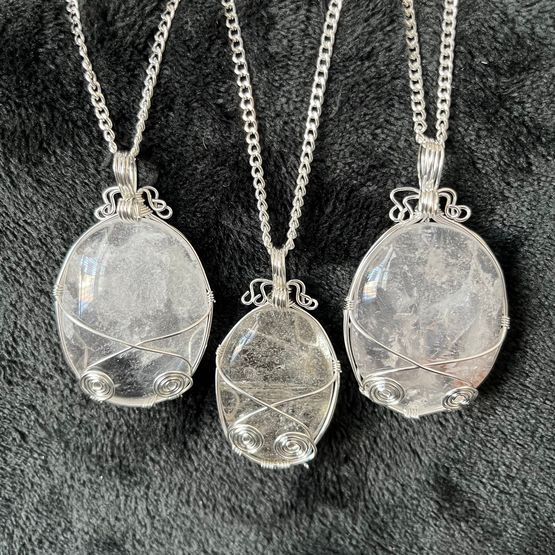 Three silver fancy wire wrapped clear quartz oval stone pendants attatched to a silver chains, displayed against a black background