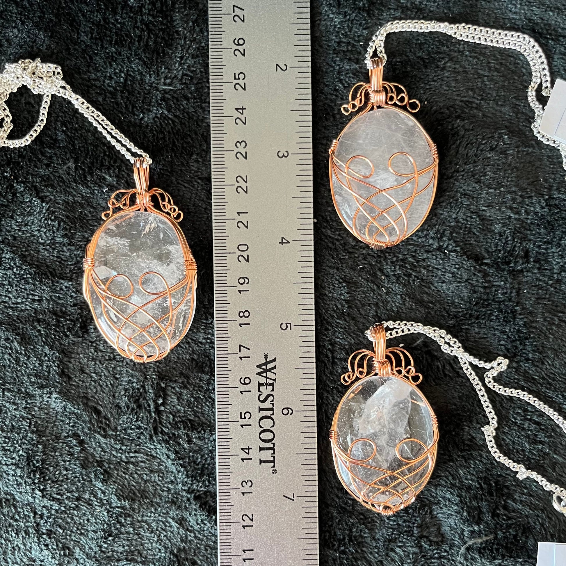 Three copper fancy wire wrapped clear quartz oval stone pendants attatched to a silver chains, displayed against a black background next to a ruler.  eaxh oendant is 1 3/4" -  2 1/4" long