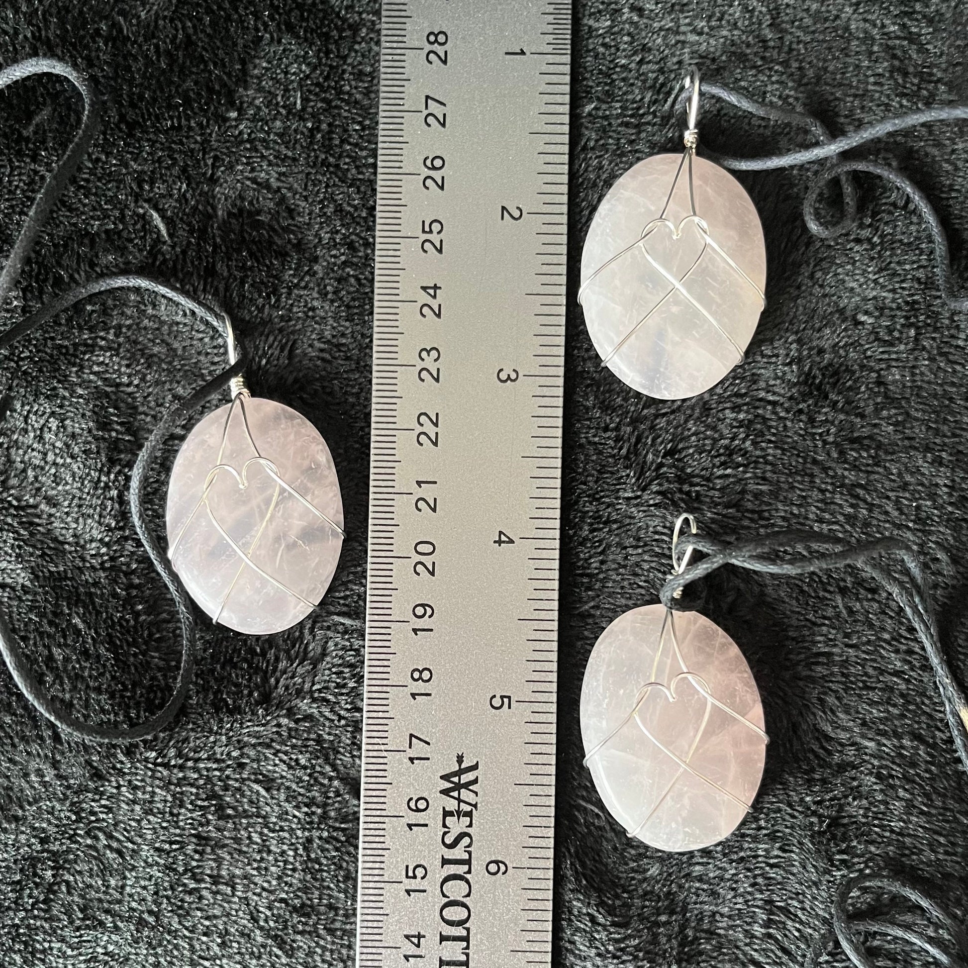 3 ornately silver wire wrapped translicent pink rose quartz worry stone pendants, approximately 1 1/4" long, attarched to adjustable black cords, displayed next to a ruler. The ailver wire male a heart shape in the center pf the rose quartz stone.